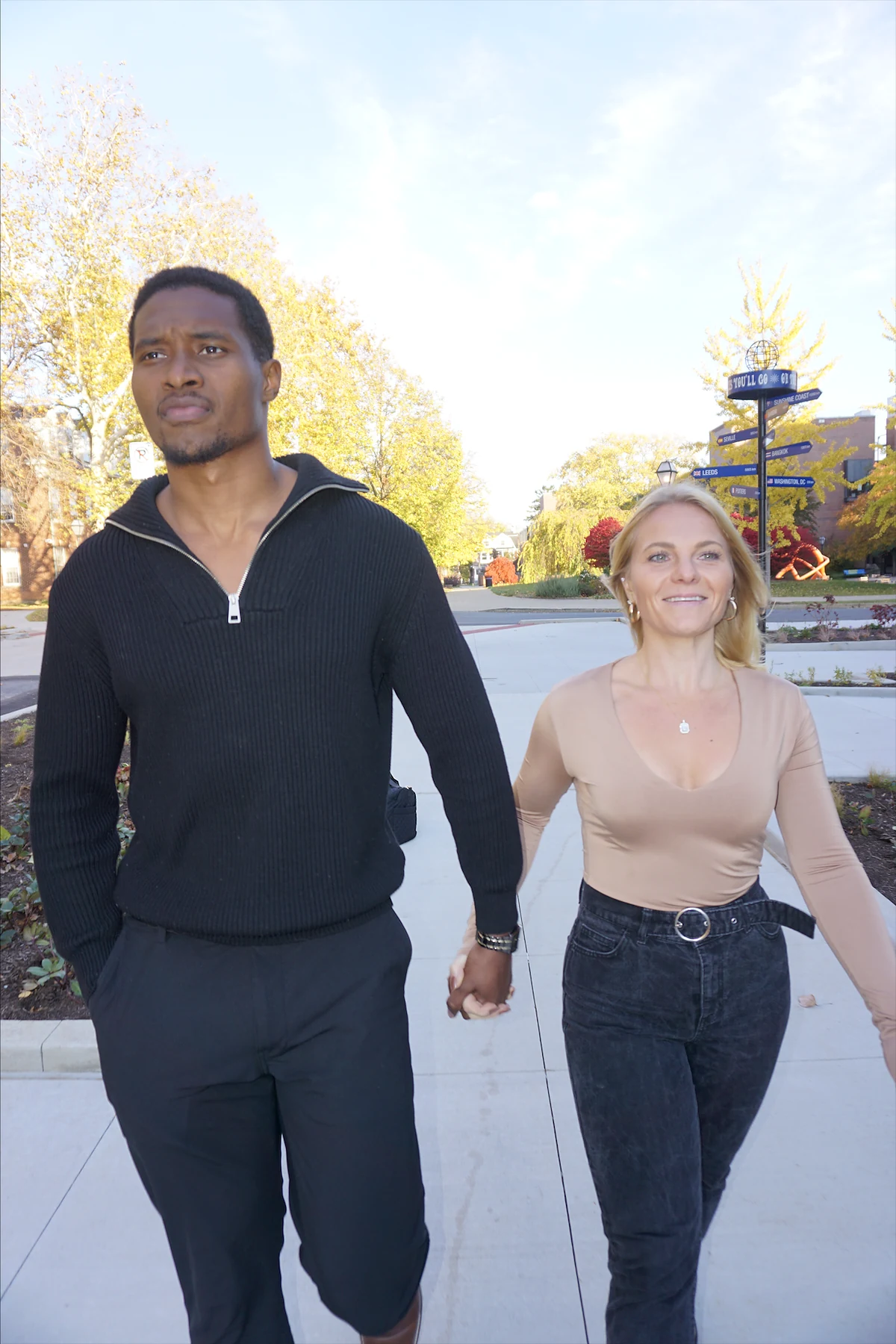 Lacey Cook and Iván Quintero, founders of Move With Intention, walk toward the camera hand in hand, smiling, on a path lined with autumn trees.